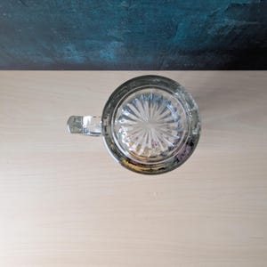 K&ouml;nnte beinhalten: Klarer Bierkrug aus Glas mit Henkel, von oben betrachtet. Der Boden des Krugs weist ein Sternmuster auf. Der Krug steht auf einer hellen Holzoberfl&auml;che, mit einem dunkel t&uuml;rkis-blauen Hintergrund.