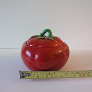 May include: A ceramic tomato-shaped container with a lid. The container is a glossy red with a green stem and leaf detail. A measuring tape shows the container is approximately 10 cm wide.