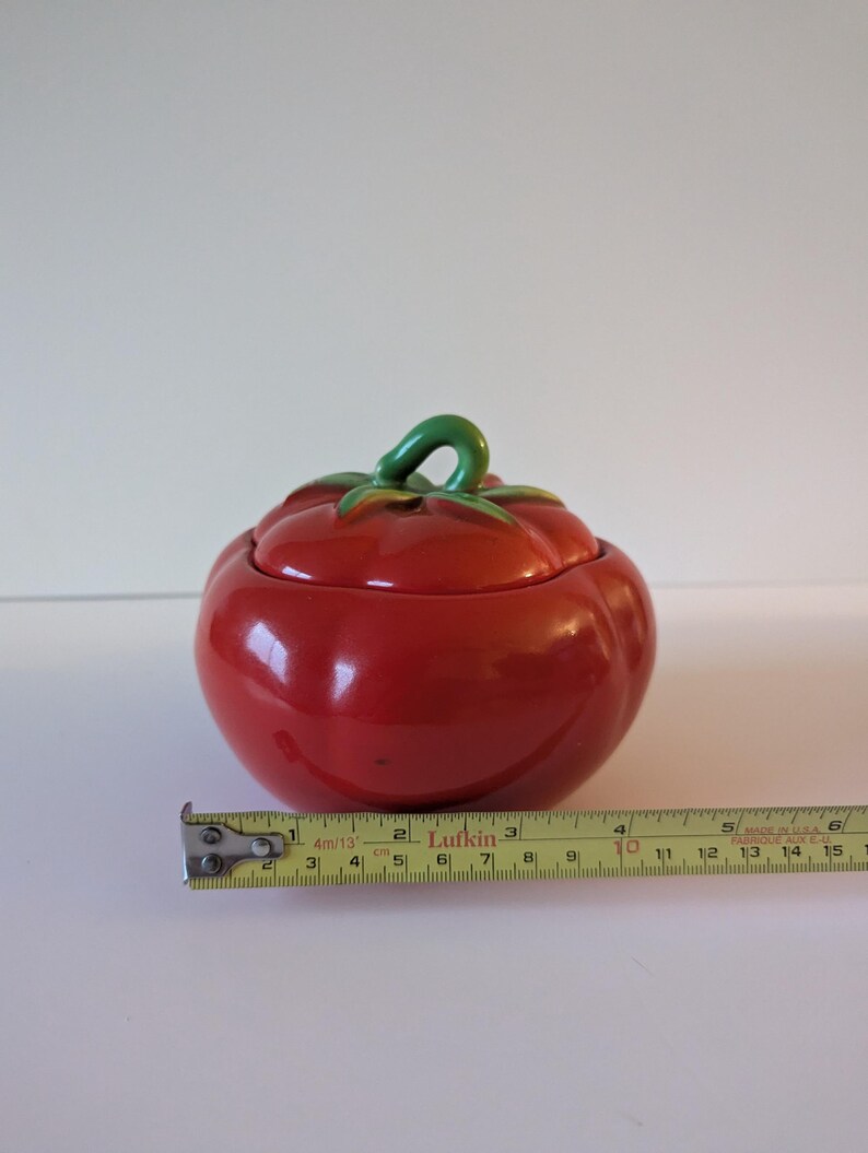May include: A ceramic tomato-shaped container with a lid. The container is a glossy red with a green stem and leaf detail. A measuring tape shows the container is approximately 10 cm wide.