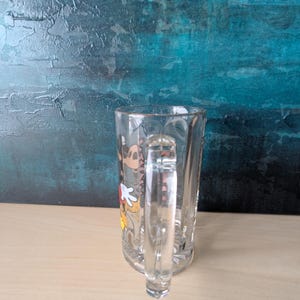 K&ouml;nnte beinhalten: Klarer Bierkrug aus Glas mit Henkel. Der Krug zeigt eine farbenfrohe Grafik von Mickey Mouse. Der Krug ist leer und steht auf einer hellbraunen Oberfl&auml;che, mit einem strukturierten t&uuml;rkisblauen und blauen Hintergrund.