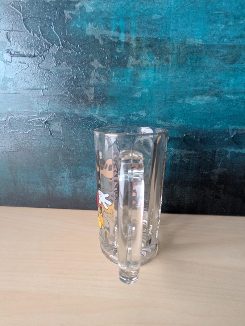 K&ouml;nnte beinhalten: Klarer Bierkrug aus Glas mit Henkel. Der Krug zeigt eine farbenfrohe Grafik von Mickey Mouse. Der Krug ist leer und steht auf einer hellbraunen Oberfl&auml;che, mit einem strukturierten t&uuml;rkisblauen und blauen Hintergrund.