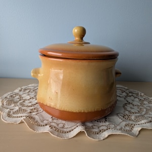 Peut inclure: Une marmite en céramique avec un corps rond et deux poignées latérales. Le pot est émaillé dans des tons de jaune et de marron. Le pot est posé sur un napperon en dentelle blanche.