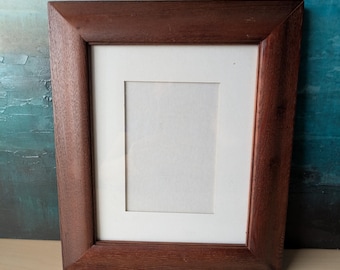 May include: A rectangular, dark brown wooden picture frame with a white mat. The frame has a beveled edge and a smooth finish. The background is a textured blue and green abstract painting.