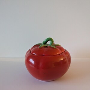 May include: A red ceramic tomato-shaped container with a lid. The container has a glossy finish and a green stem detail. The lid fits neatly, and the design is a charming kitchen accessory, perfect for storing small items.