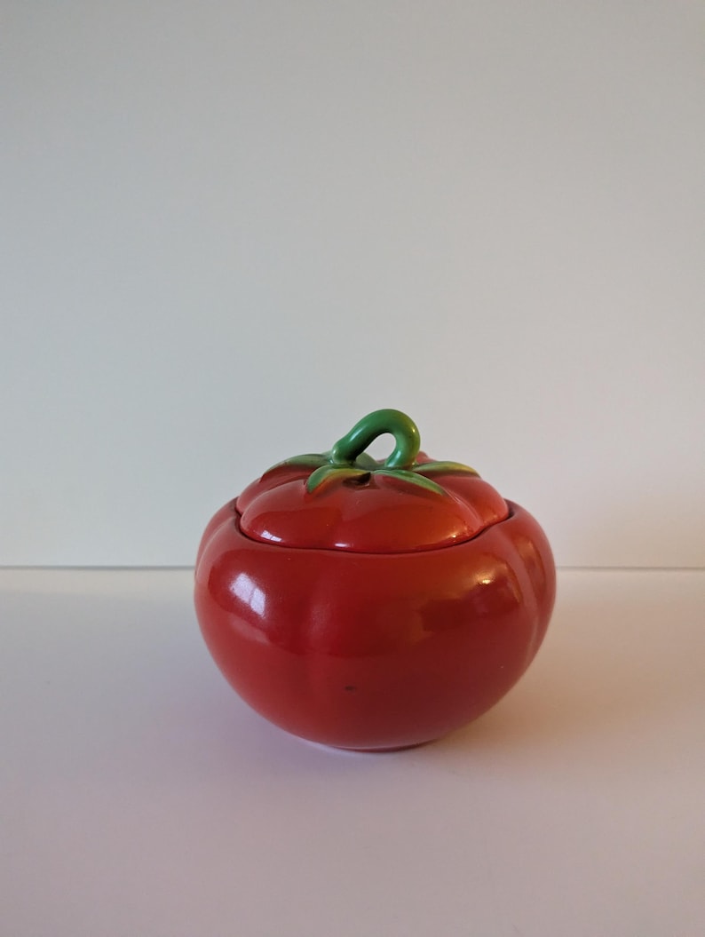 May include: A red ceramic tomato-shaped container with a lid. The container has a glossy finish and a green stem detail. The lid fits neatly, and the design is a charming kitchen accessory, perfect for storing small items.
