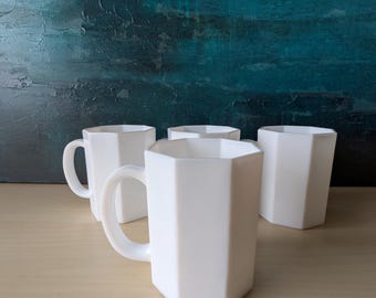 May include: Four white, geometric-shaped mugs with handles are arranged on a light-colored wooden surface. The mugs have a modern, minimalist design with a faceted exterior. The background features a textured blue and teal abstract painting.