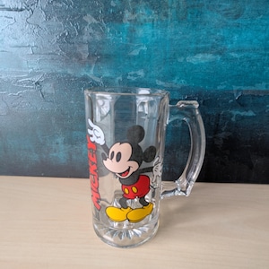 K&ouml;nnte beinhalten: Klarer Bierkrug aus Glas mit Henkel, verziert mit einem farbenfrohen Bild von Mickey Mouse. Die Zeichentrickfigur tr&auml;gt rote Shorts, gelbe Schuhe und wei&szlig;e Handschuhe. Das Wort "Mickey" ist rot auf der Seite des Bechers aufgedruckt.