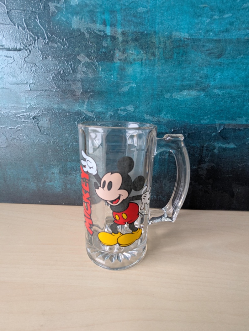 K&ouml;nnte beinhalten: Klarer Bierkrug aus Glas mit Henkel, verziert mit einem farbenfrohen Bild von Mickey Mouse. Die Zeichentrickfigur tr&auml;gt rote Shorts, gelbe Schuhe und wei&szlig;e Handschuhe. Das Wort "Mickey" ist rot auf der Seite des Bechers aufgedruckt.