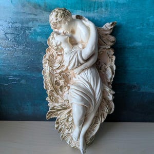 May include: A cream-coloured wall sculpture of a couple embracing. The figures are in classical attire, with the man leaning over the woman. The sculpture is set against a decorative, leaf-shaped background with gold accents.