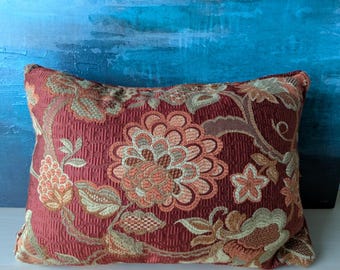 May include: Rectangular decorative pillow with a floral pattern. The pillow features a deep red background with a repeating pattern and large flowers in shades of peach, gold, and green. The pillow is placed on a white surface against a blue background.