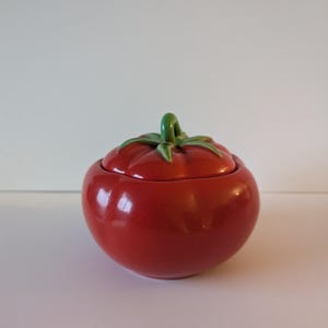 May include: A ceramic tomato-shaped jar with a lid. The jar is a glossy red with a green stem and leaves on top. The jar is designed to resemble a ripe tomato.