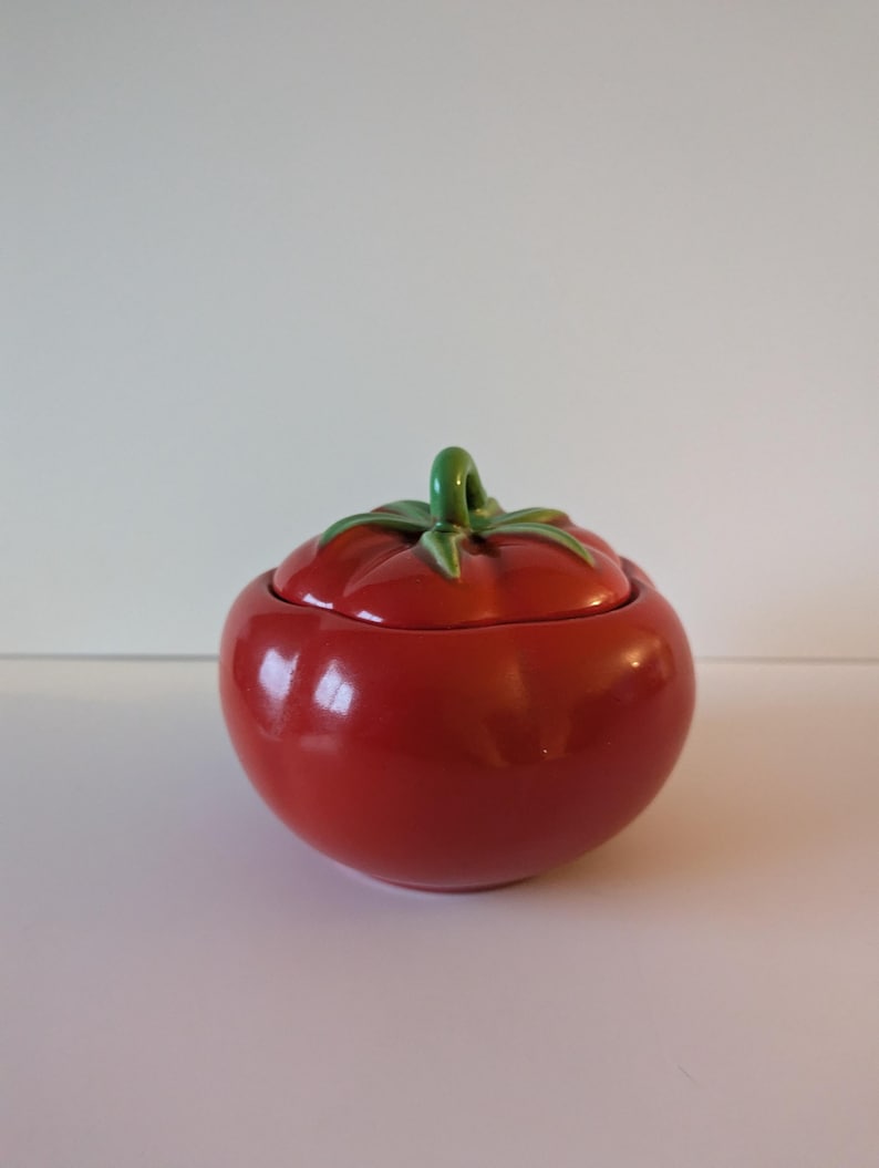 May include: A ceramic tomato-shaped jar with a lid. The jar is a glossy red with a green stem and leaves on top. The jar is designed to resemble a ripe tomato.