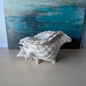 May include: A ceramic seashell-shaped bowl with a pearlescent finish. The bowl has a textured surface and stands on three small feet. The background features an abstract painting in shades of blue, teal, and grey.