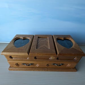 Puede incluir: Una caja de joyería de madera con un acabado marrón claro. La caja tiene tres secciones en la parte superior, dos con inserciones de vidrio en forma de corazón. Dos pequeños cajones con asas de metal son visibles debajo. La caja se encuentra sobre una superficie clara.
