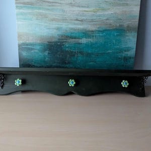 May include: A dark green wooden shelf with a scalloped bottom edge. The shelf has three flower-shaped knobs with blue and yellow accents. Decorative black metal brackets support the shelf. The shelf is displayed against a blue and teal abstract painting.