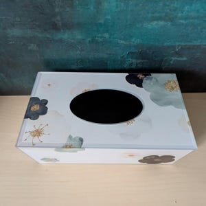 Puede incluir: Una caja para pañuelos rectangular con base blanca decorada con motivos florales estilo acuarela en tonos azul oscuro, verde salvia y rosa pálido, acentuada con pequeños detalles de purpurina dorada alrededor de una abertura ovalada.