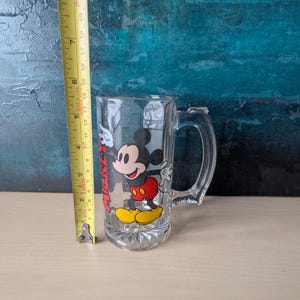 K&ouml;nnte beinhalten: Klarer Bierkrug aus Glas mit Mickey Mouse. Der Krug hat einen Henkel und ein gedrucktes Bild von Mickey Mouse in seinem klassischen Outfit. Der Krug ist etwa 15 cm hoch.