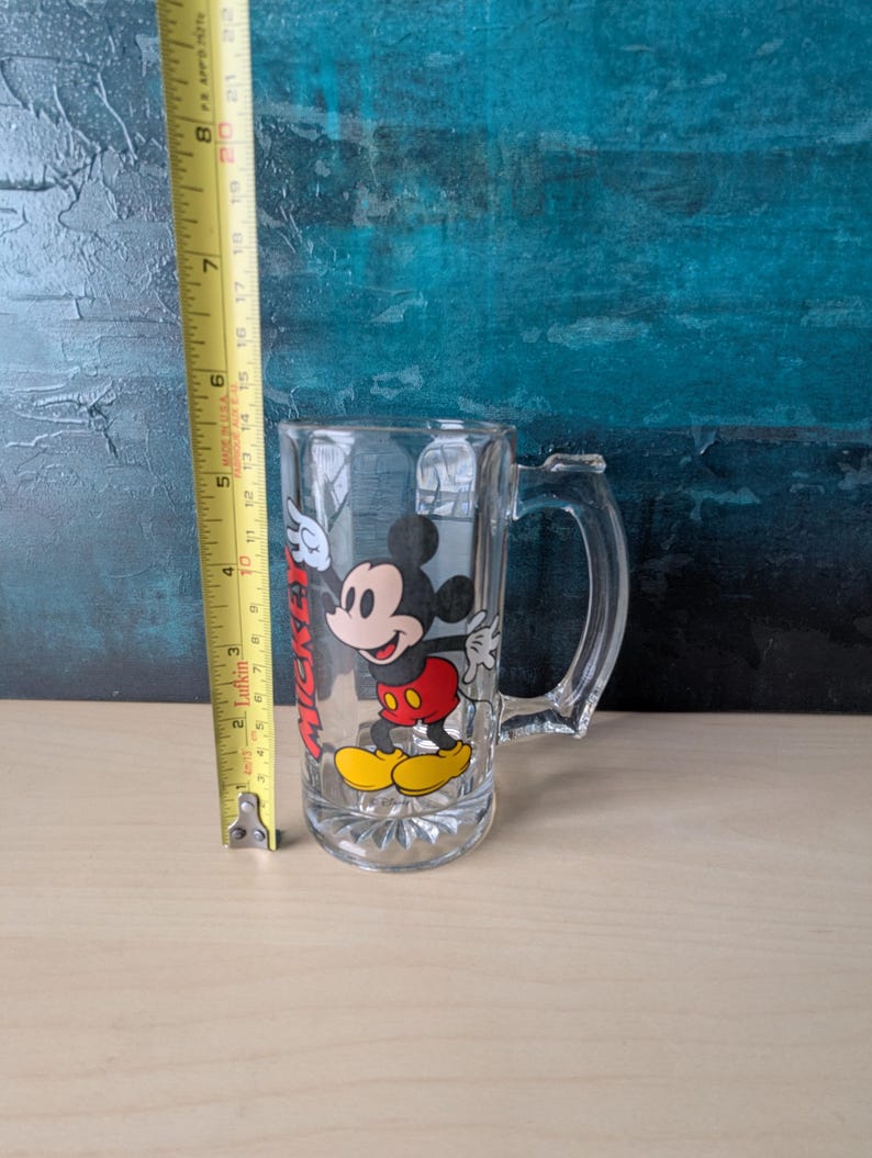 K&ouml;nnte beinhalten: Klarer Bierkrug aus Glas mit Mickey Mouse. Der Krug hat einen Henkel und ein gedrucktes Bild von Mickey Mouse in seinem klassischen Outfit. Der Krug ist etwa 15 cm hoch.