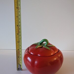 May include: A ceramic tomato-shaped jar with a lid. The jar is a vibrant red with a glossy finish. The lid features green leaves and a green stem. The jar measures approximately 15 cm in height.