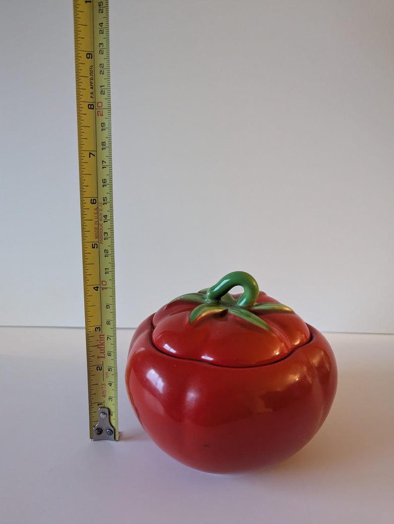 May include: A ceramic tomato-shaped jar with a lid. The jar is a vibrant red with a glossy finish. The lid features green leaves and a green stem. The jar measures approximately 15 cm in height.