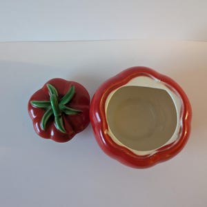May include: A ceramic tomato-shaped cookie jar with a matching lid. The jar is bright red with a white interior and a green stem on the lid. The lid is separate from the jar, showing the inside of the jar.