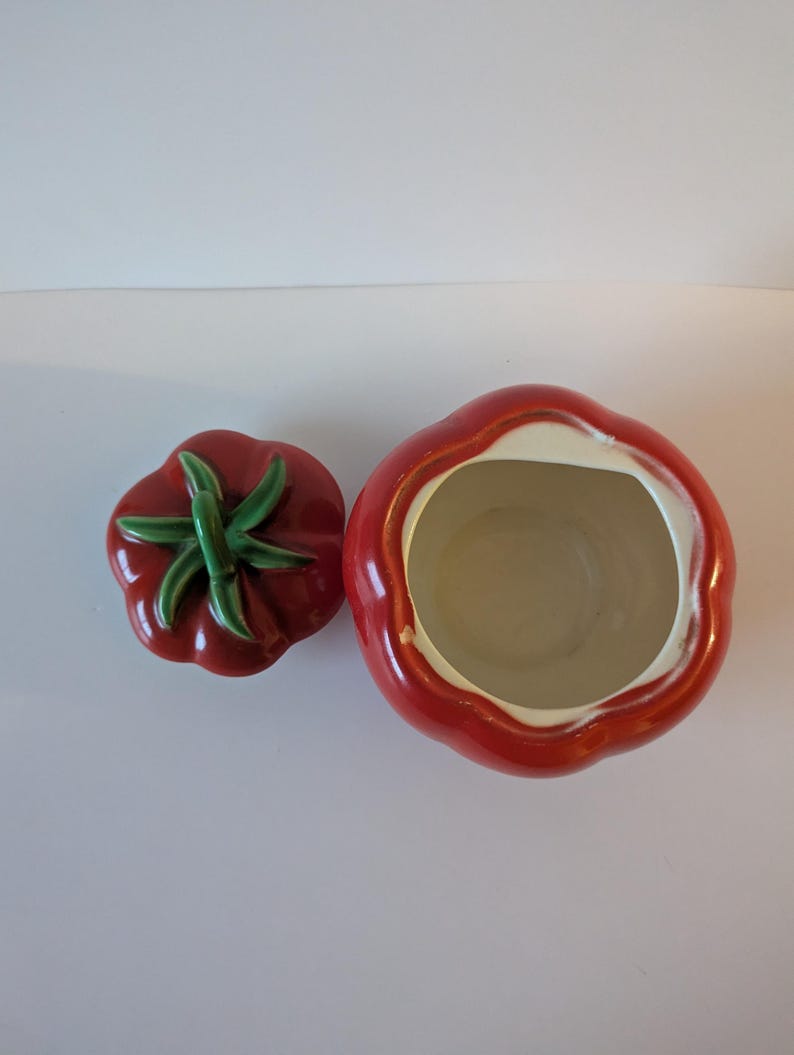 May include: A ceramic tomato-shaped cookie jar with a matching lid. The jar is bright red with a white interior and a green stem on the lid. The lid is separate from the jar, showing the inside of the jar.