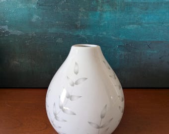 May include: A white ceramic vase with a teardrop shape. The vase features a pattern of gray leaves and stems. The vase is sitting on a wooden surface, with a blue and green abstract painting in the background.