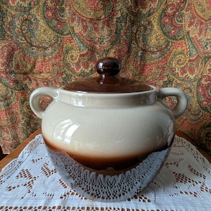 Puede incluir: Una fuente de cerámica con tapa. El plato es blanquecino con una banda marrón alrededor del centro y una tapa marrón. Tiene dos asas y se asienta sobre un tapete de encaje blanco. El fondo es una tela con estampado de cachemira.