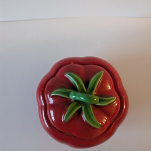 May include: A ceramic tomato-shaped container with a lid. The container is a glossy red with a green stem and leaves on top. The design is a whimsical, decorative piece.