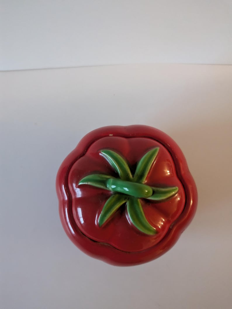 May include: A ceramic tomato-shaped container with a lid. The container is a glossy red with a green stem and leaves on top. The design is a whimsical, decorative piece.