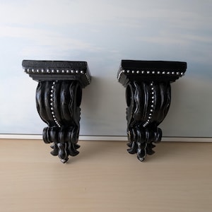 Op de afbeelding: Twee zwarte decoratieve wandplanken met sierlijke details. Elke plank heeft een vierkante bovenkant met zilveren kraalaccenten, een gebogen steun en een decoratieve onderkant. De planken worden gepresenteerd tegen een lichtblauwe en witte achtergrond.
