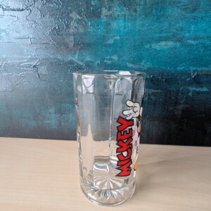 K&ouml;nnte beinhalten: Klares Glas mit dem Wort "MICKEY" in Rot, einer wei&szlig;en behandschuhten Hand und Zeichentrickfiguren. Das Glas hat einen facettierten Boden und einen geraden, zylindrischen K&ouml;rper. Das Design ist ein klassisches Cartoon-Thema.
