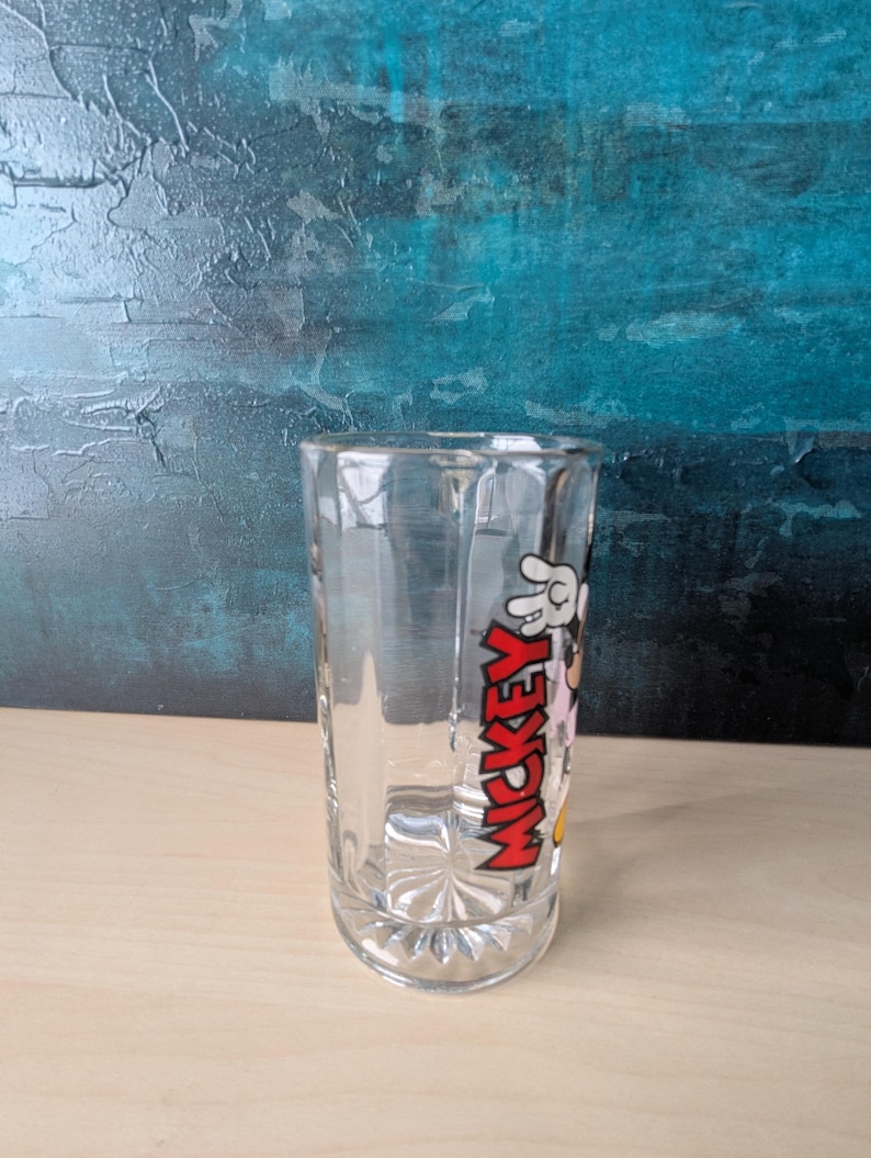 K&ouml;nnte beinhalten: Klares Glas mit dem Wort "MICKEY" in Rot, einer wei&szlig;en behandschuhten Hand und Zeichentrickfiguren. Das Glas hat einen facettierten Boden und einen geraden, zylindrischen K&ouml;rper. Das Design ist ein klassisches Cartoon-Thema.