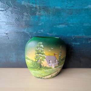 May include: A green ceramic vase featuring a hand-painted landscape. The artwork shows a house with a brown roof, a tree, and a yellow and green field. The vase is on a wooden surface against a blue backdrop.