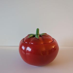 May include: A ceramic tomato-shaped container with a lid. The container is a glossy red with a green stem and leaves on top. The lid fits snugly on the container, creating a complete tomato shape.