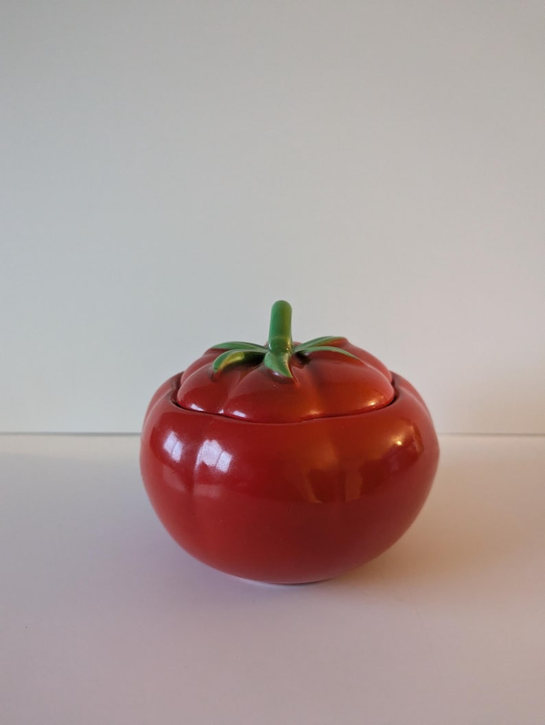 May include: A ceramic tomato-shaped container with a lid. The container is a glossy red with a green stem and leaves on top. The lid fits snugly on the container, creating a complete tomato shape.