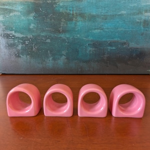 Może przedstawiać: Cztery różowe pierścienie na serwetki o zaokrąglonym kwadratowym kształcie. Ułożone w rzędzie na drewnianej powierzchni. Tło przedstawia abstrakcyjny obraz w odcieniach niebieskiego i zielonego.