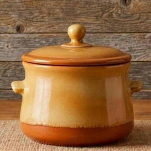 May include: A round ceramic pot with a lid, featuring a two-tone glaze with a warm honey-yellow upper section and a darker terracotta-orange base. The pot includes two small side handles and a central knob on the lid for easy lifting and handling.