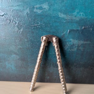 Pode incluir: Um quebra-nozes prateado com cabos texturizados e um design articulado. O quebra-nozes está aberto, pronto para uso, contra um fundo azul e turquesa.