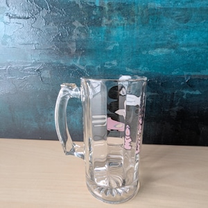 K&ouml;nnte beinhalten: Ein klarer Bierkrug mit Henkel. Der Krug hat eine rechteckige Form mit vertikalen Rippen und einem Design in Rosa und Schwarz. Der Hintergrund ist ein strukturiertes T&uuml;rkis und Blau.