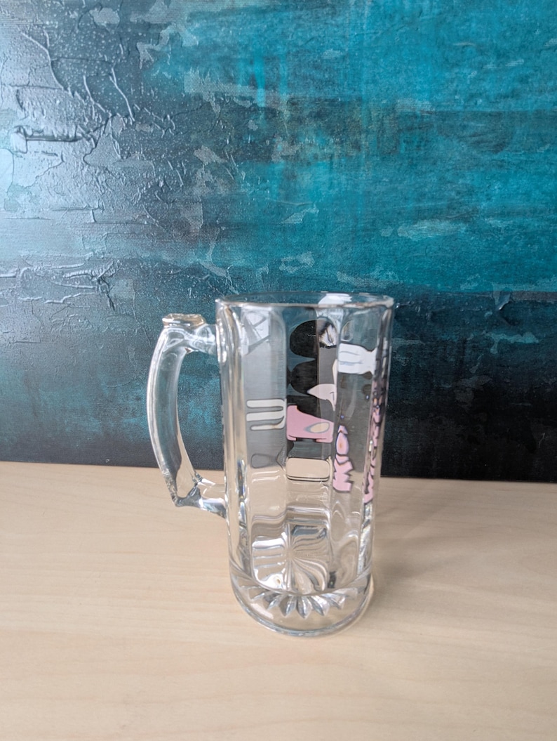 K&ouml;nnte beinhalten: Ein klarer Bierkrug mit Henkel. Der Krug hat eine rechteckige Form mit vertikalen Rippen und einem Design in Rosa und Schwarz. Der Hintergrund ist ein strukturiertes T&uuml;rkis und Blau.