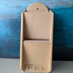 May include: A light-coloured wooden wall organiser featuring two stacked open compartments for mail or documents and three small metal hooks at the bottom for hanging items. The unit has a rounded top edge with a pre-drilled hole for mounting.