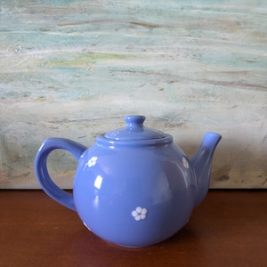 May include: A periwinkle blue ceramic teapot with a matching lid. The teapot features a rounded body, a curved handle, and a spout. White floral designs are scattered across the surface. The teapot is sitting on a brown surface.