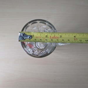 K&ouml;nnte beinhalten: Ein klares Trinkglas aus geschliffenem Glas mit einem dekorativen Muster. Das Glas wird mit einem gelben Ma&szlig;band gemessen, das einen Durchmesser von etwa 7,6 cm anzeigt.