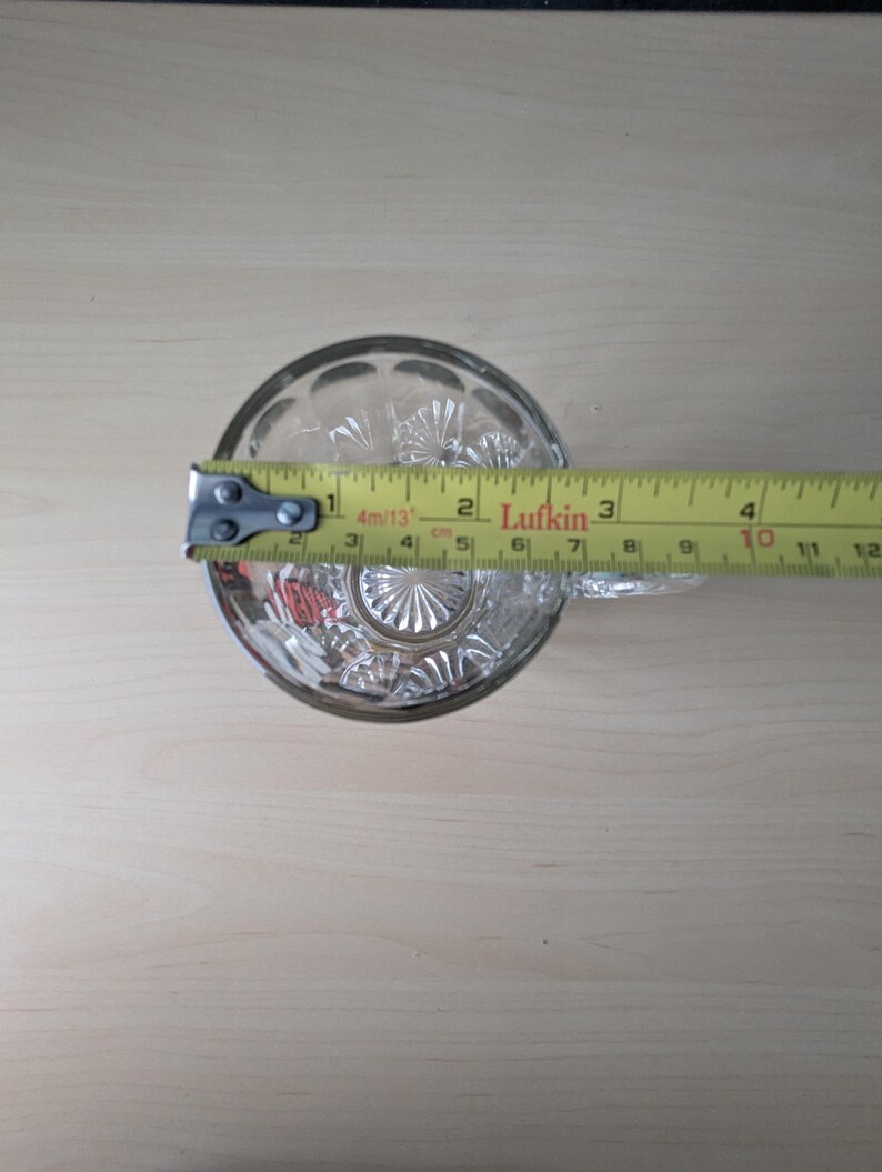 K&ouml;nnte beinhalten: Ein klares Trinkglas aus geschliffenem Glas mit einem dekorativen Muster. Das Glas wird mit einem gelben Ma&szlig;band gemessen, das einen Durchmesser von etwa 7,6 cm anzeigt.