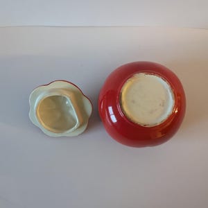May include: A red ceramic jar with a matching lid. The jar is round with a glossy finish. The lid is flower-shaped with a white interior and red trim. The jar and lid are on a white surface.