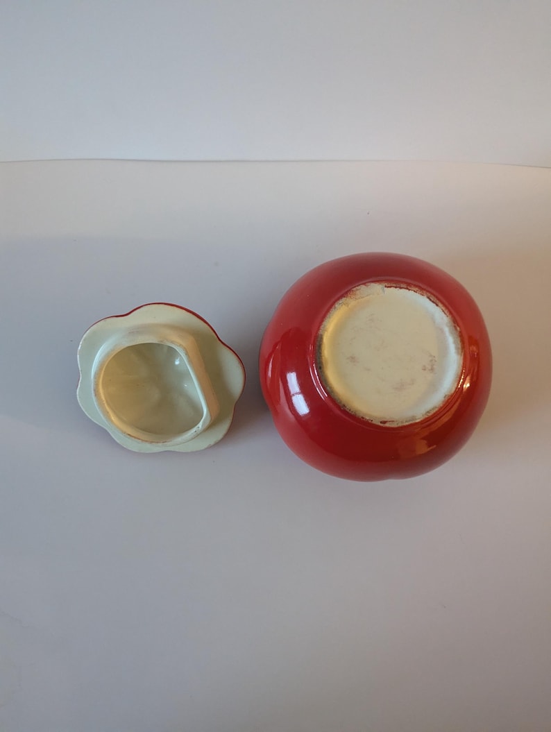 May include: A red ceramic jar with a matching lid. The jar is round with a glossy finish. The lid is flower-shaped with a white interior and red trim. The jar and lid are on a white surface.
