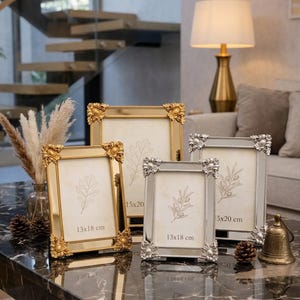 Könnte beinhalten: Vier dekorative Bilderrahmen in Gold und Silber mit aufwendigen Eckdetails. Zwei Rahmen sind goldfarben und zwei silberfarben. Die Rahmen zeigen botanische Illustrationen und sind 13x18 cm und 15x20 cm groß.