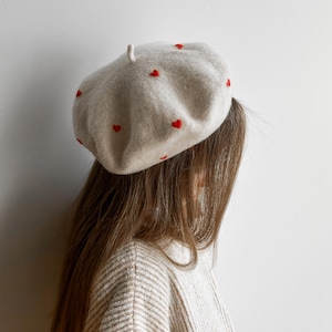 Può includere: Un basco in feltro beige con piccoli cuori rossi ricamati sparsi sulla superficie. Il cappello presenta una corona arrotondata con una piccola linguetta in tessuto centrata sulla parte superiore, offrendo un aspetto morbido e strutturato.