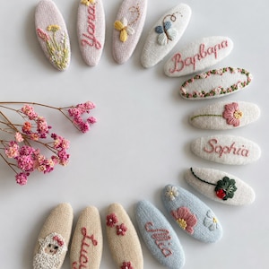 May include: A collection of oval-shaped hair clips featuring embroidered designs on textured fabric. The clips display various motifs including flowers, a sheep, and names such as Yana, Barbara, Sophia, and Mia, arranged in a circular pattern.